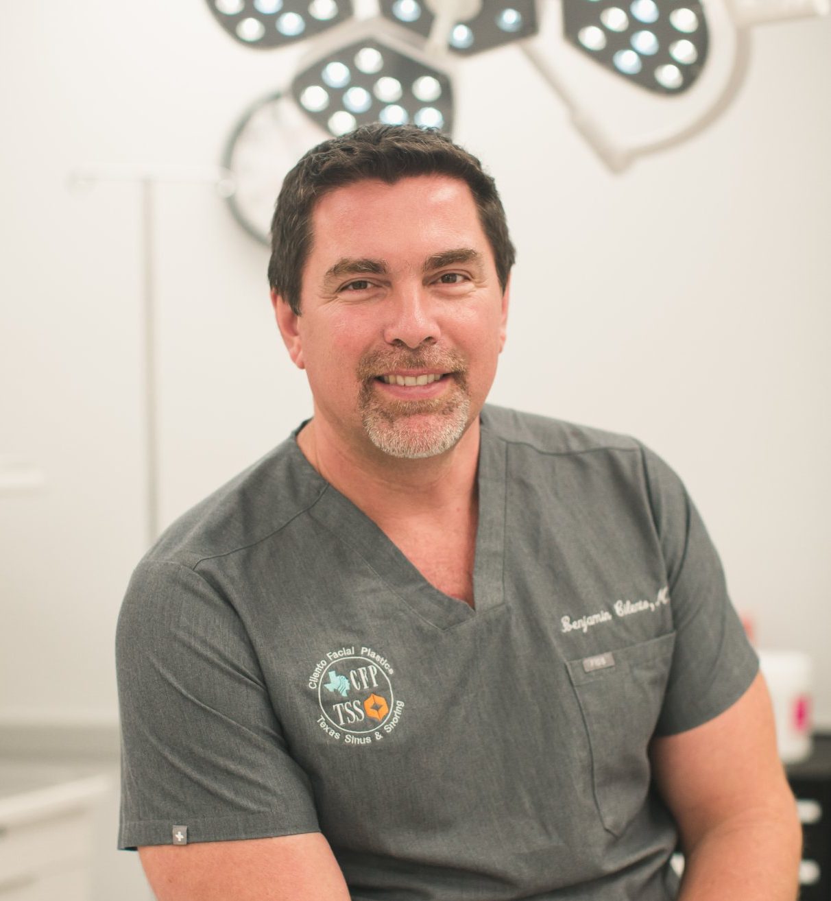 Dr. Ben Cilento smiling in surgical scrubs in an operating room with overhead surgical lights